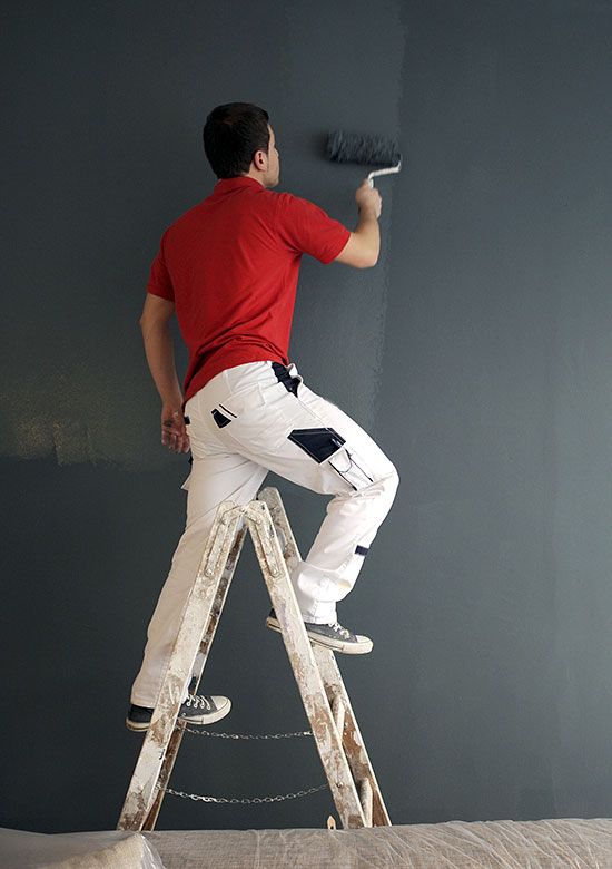 Ein Maler steht auf einer Leiter und streicht eine dunkle Wand mit der Rolle.