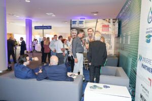 Besucher stehen und sprechen an Messeständen; einige sitzen auf Sofas im Vordergrund.