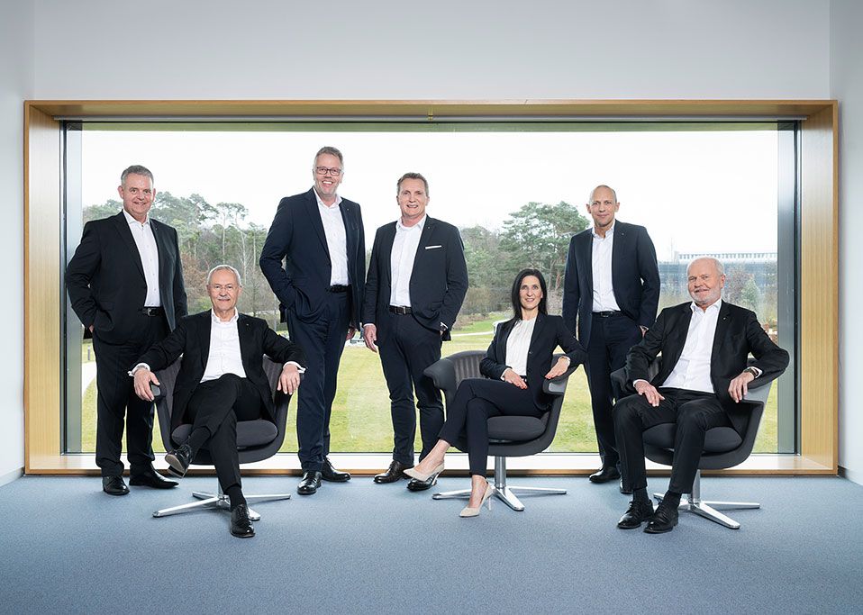 Sieben Personen in Businesskleidung posieren vor einer großen Fensterfront; drei sitzen, vier stehen.