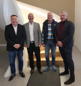 Vier Männer stehen nebeneinander und lächeln in einem hellen Innenraum.