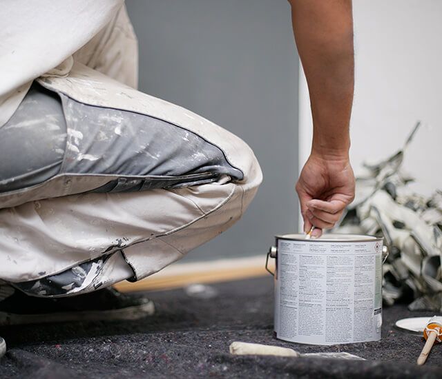 Ein Maler kniet am Boden und öffnet eine Farbdose; daneben liegt Werkzeug.